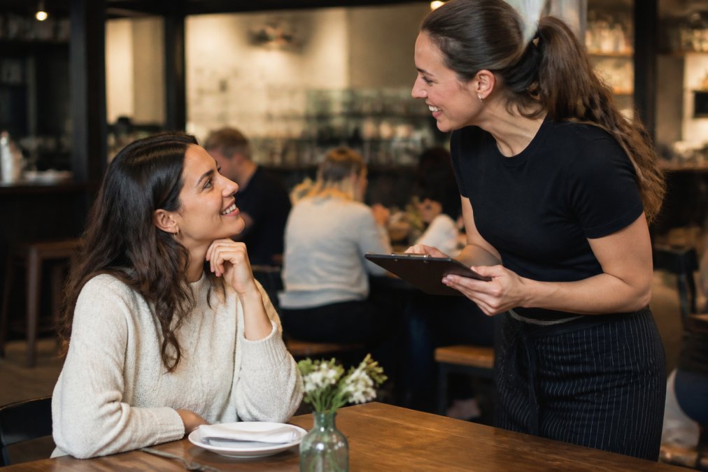A server greeting a familiar customer by name with a warm smile, recognizing them as a regular at the restaurant