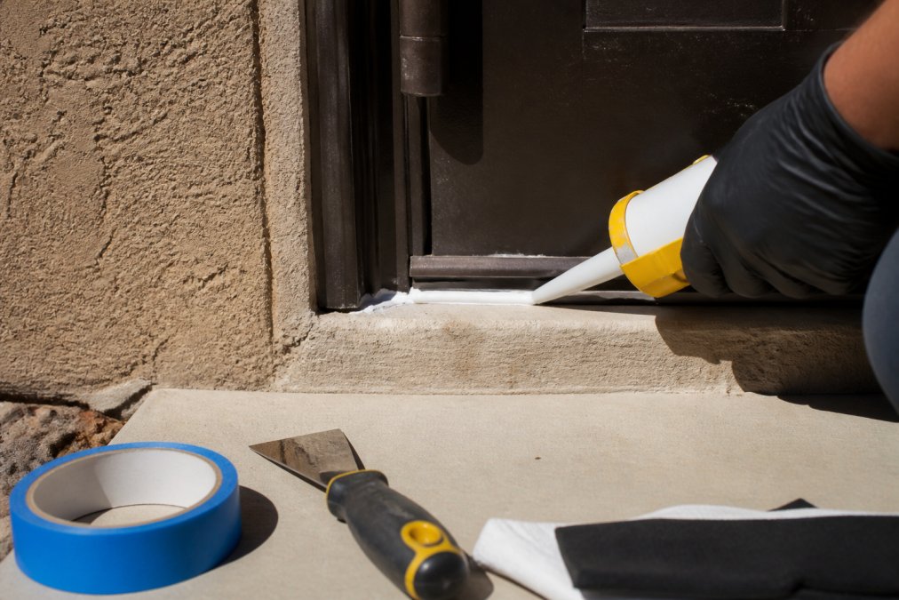 A gloved hand applying white silicone caulk along a gap at the bottom of an exterior door frame on a stucco wall, bright desert sunlight casting sharp shadows