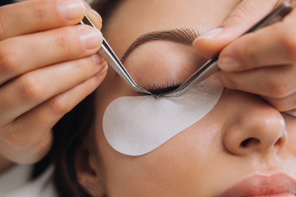 Close-up of hands using precision tweezers to apply individual lash segments to the natural lash line, demonstrating proper angle and positioning technique under bright studio lighting