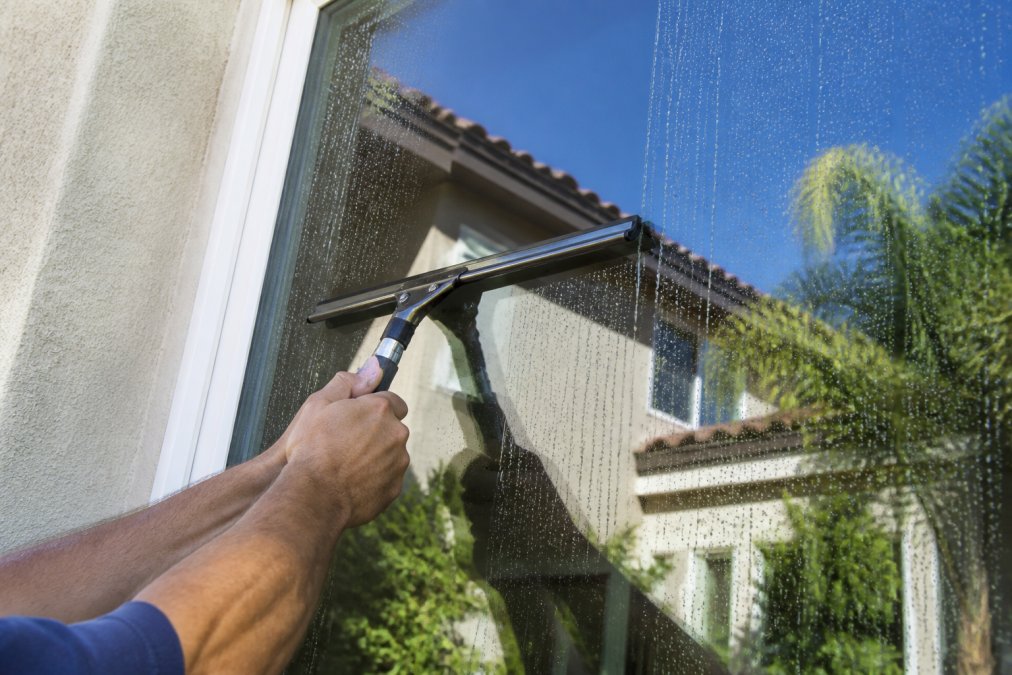Close-up of hands gripping a squeegee pulling a clean stripe down a large exterior window, water droplets beading on the glass, bright daylight, residential home exterior