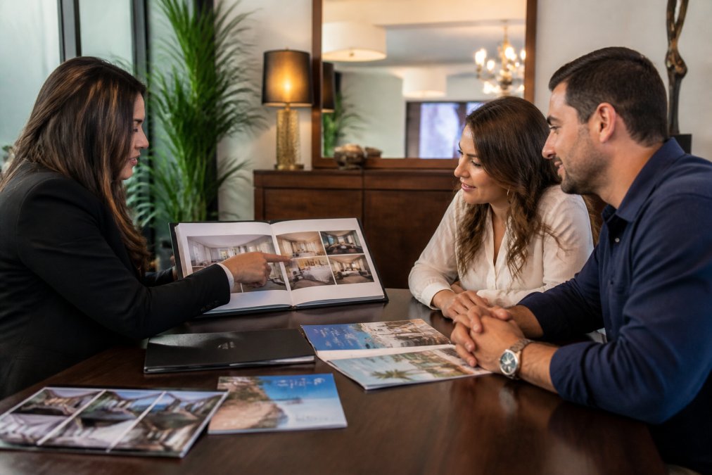 A travel advisor reviewing hotel room options on a laptop with a couple seated across the desk in a warmly lit professional office, hotel brochures spread on the table, the advisor pointing to the screen