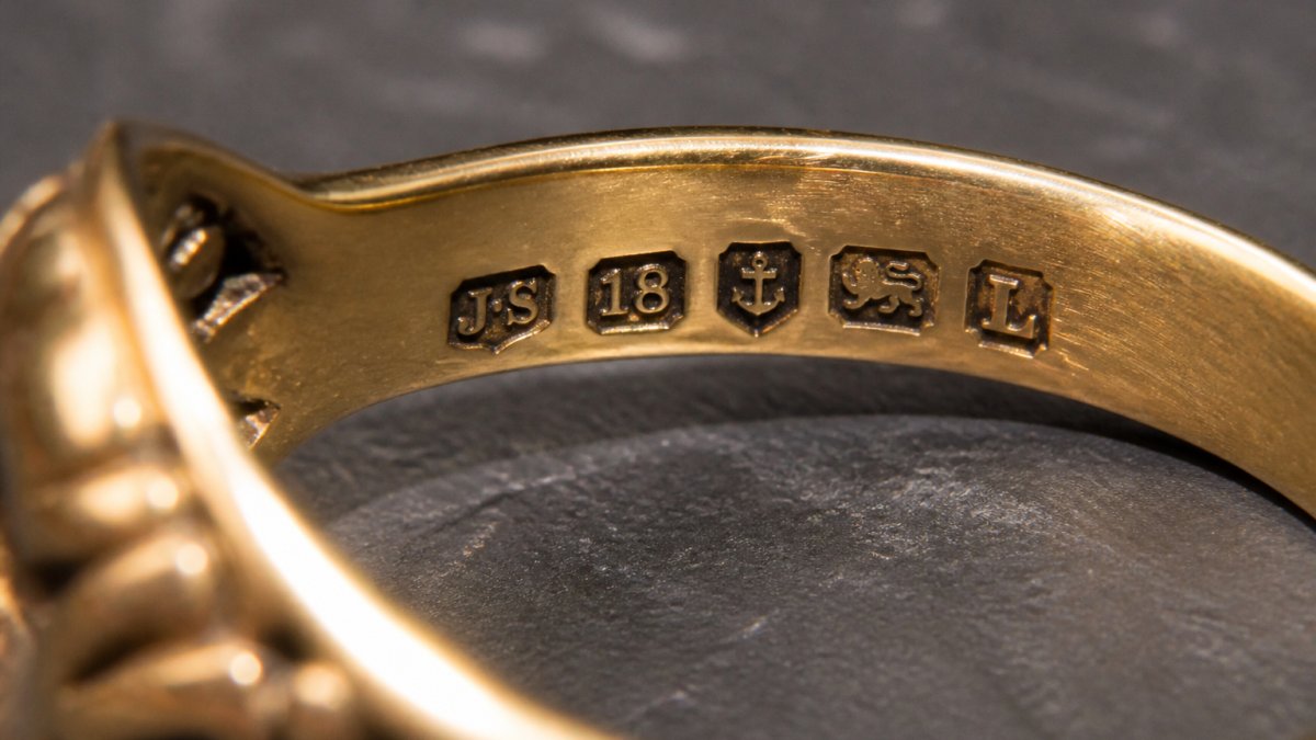 Close-up photograph of antique gold ring showing visible hallmarks and maker's marks stamped into the metal, with clear detail of the stamps under natural side lighting