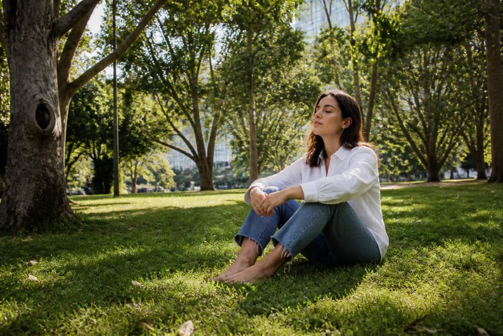How to Ground Yourself Spiritually and Reconnect 1 Person sitting barefoot on grass in a park surrounded by trees and natural greenery, appearing calm and centred with soft natural light filtering through the leaves