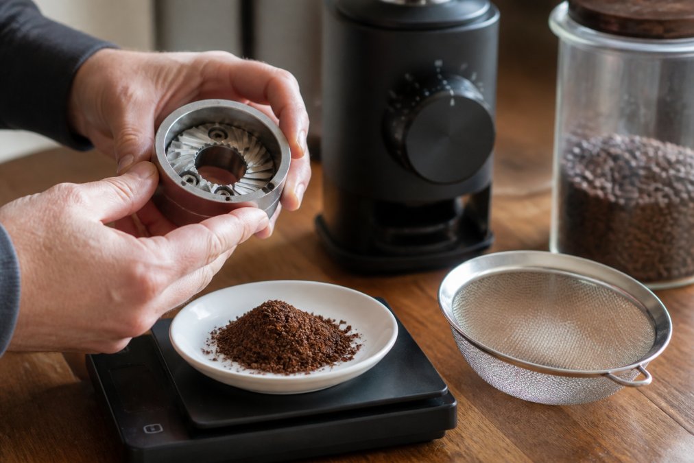 Close-up of hands holding a burr grinder burr set over a kitchen scale, with a small pile of freshly ground coffee and a sieve resting on a wooden countertop