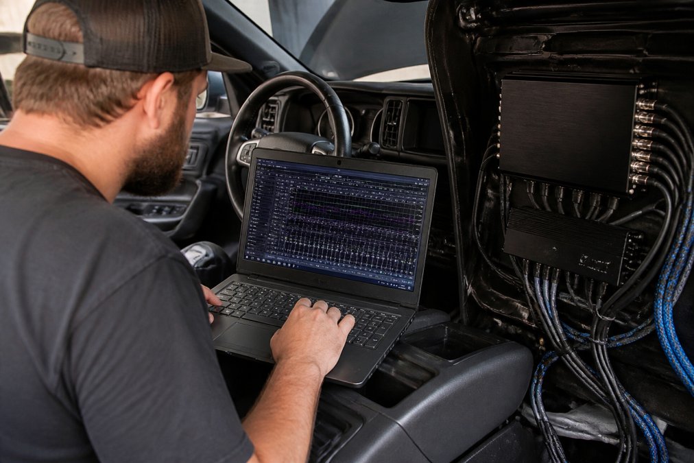A car audio technician in a workshop adjusting a DSP processor on a laptop while sitting in the driver's seat of a vehicle, with tuning software visible on the screen and audio cables running to an amplifier in the background
