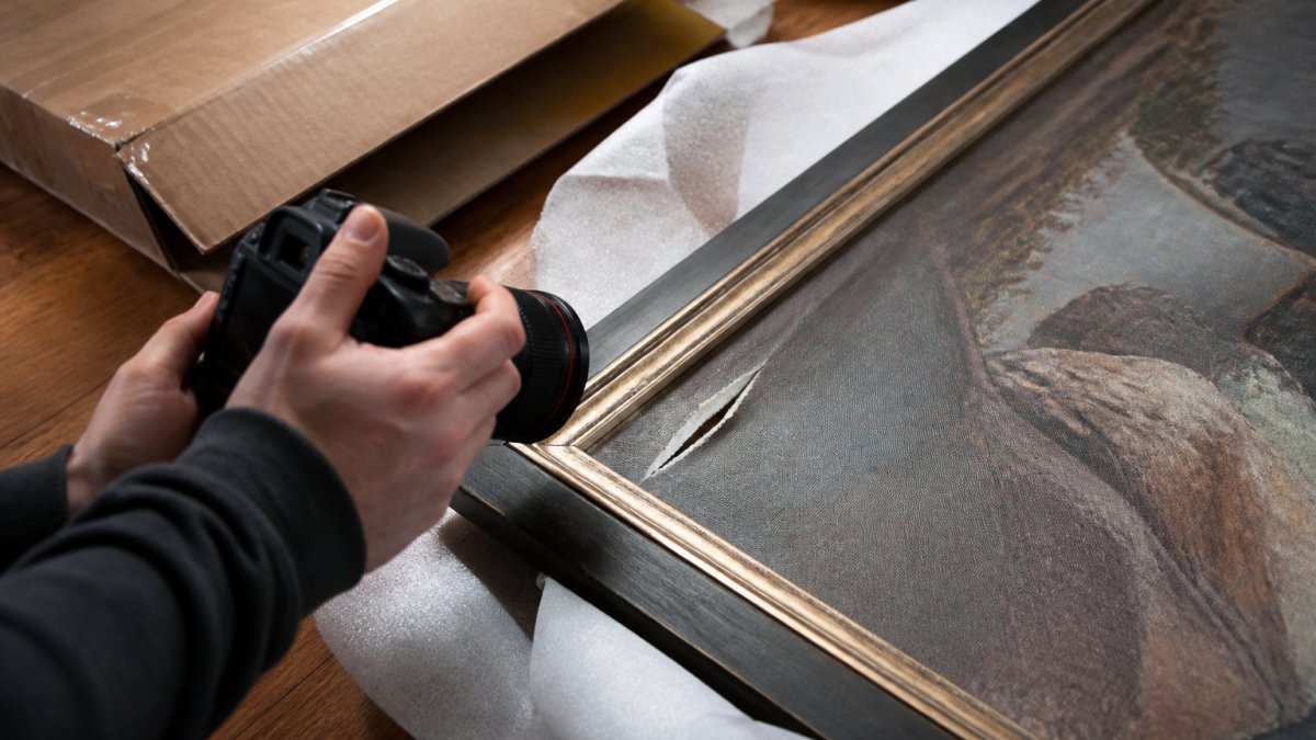 Close-up of hands photographing a framed painting with a visible tear in the canvas, with the shipping box and packing materials visible in the background on a wooden floor
