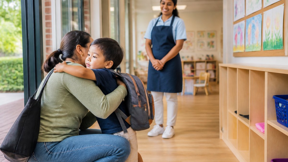 A parent kneeling to hug a toddler in a bright daycare entryway, with a teacher smiling warmly in the background and children's artwork on the walls