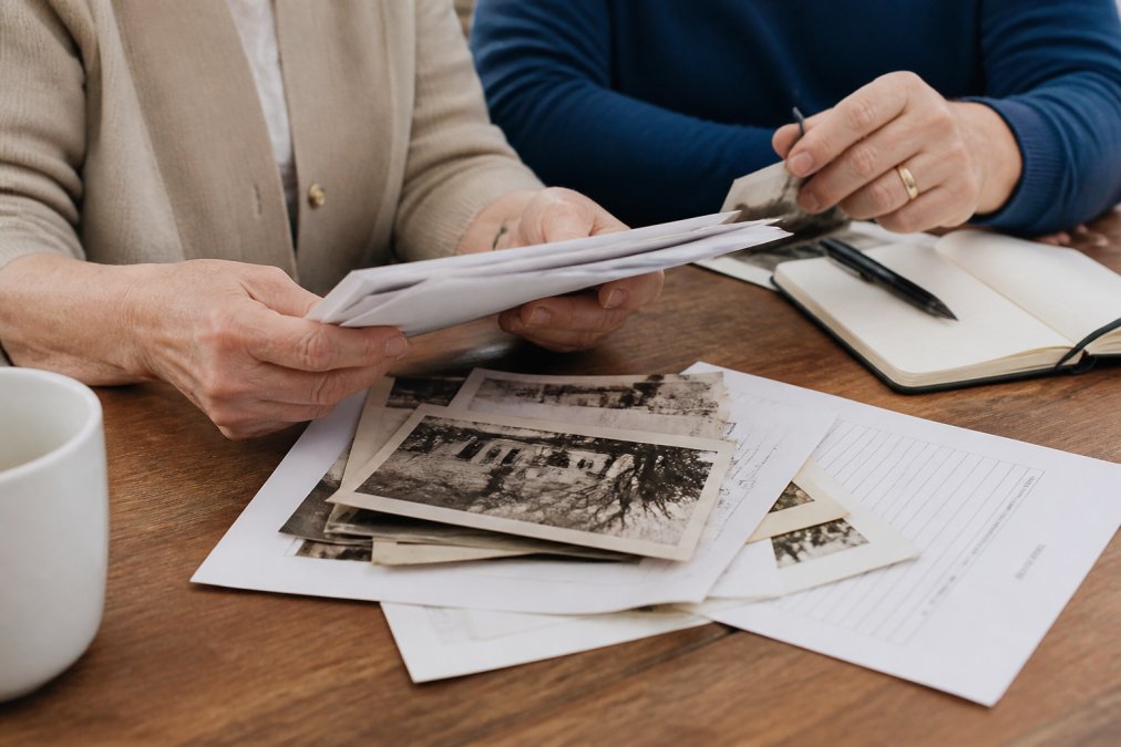 Close-up of hands holding estate documents, photos, and a checklist on a wooden table with a pen and notebook nearby