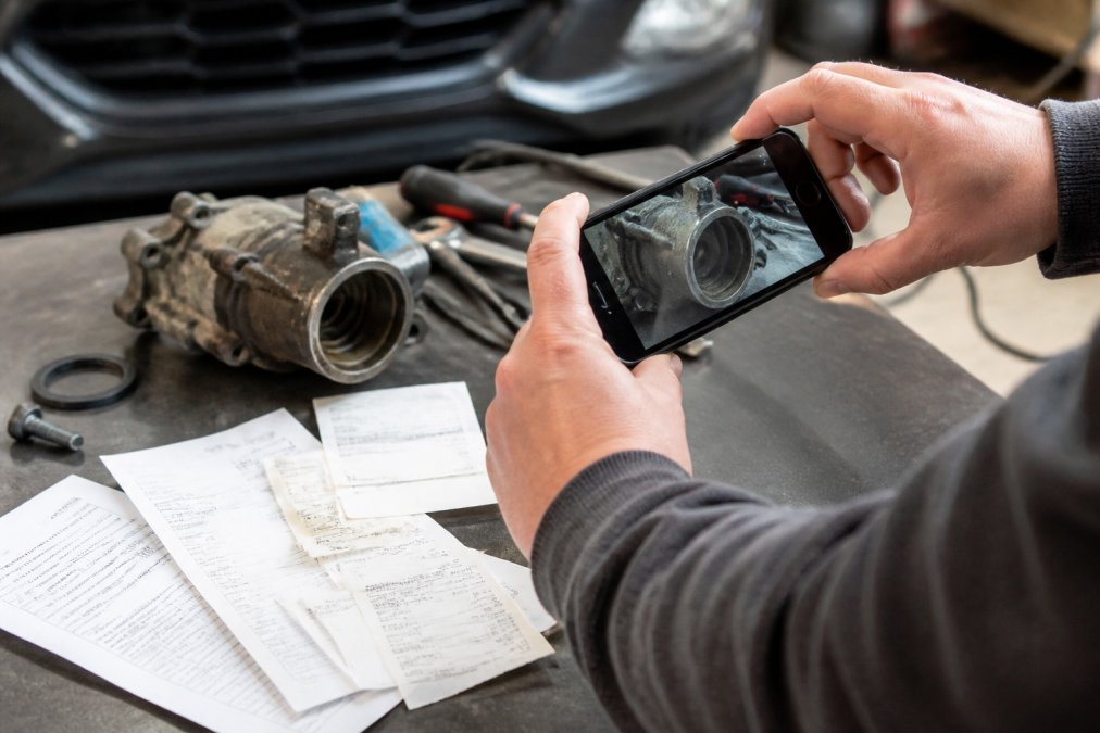 Close-up of hands photographing a damaged car component with a smartphone, with receipts and warranty documents spread across a workbench
