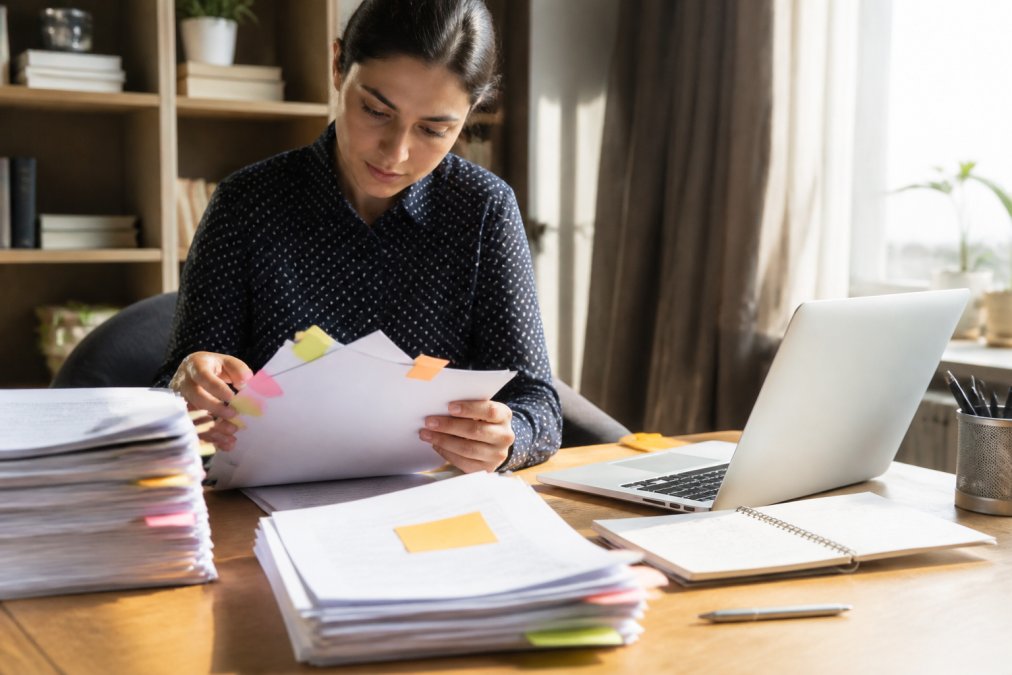 A person organizing claim paperwork at a home desk, sorting files with sticky notes while a laptop shows an open email inbox, warm natural light from a window