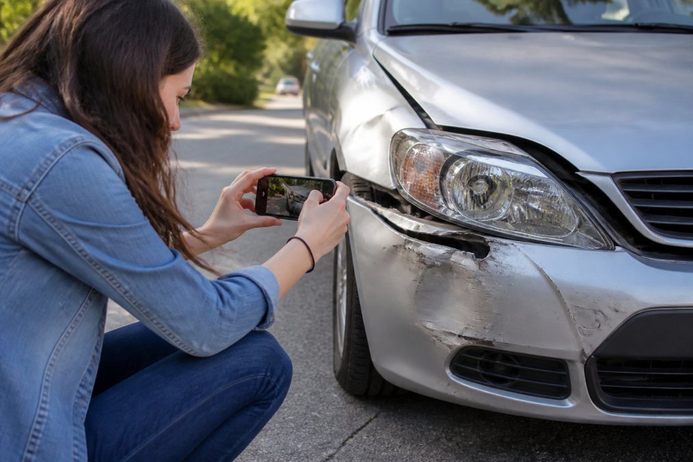 Person photographing vehicle damage at accident scene with smartphone, capturing close-up of dented bumper and broken headlight in natural daylight