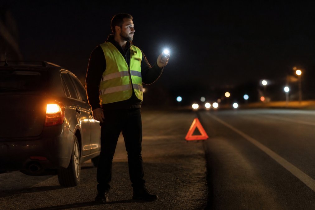 Driver standing outside vehicle at night on shoulder of road with hazard lights activated, wearing reflective vest, holding smartphone with flashlight to check surroundings, with warning triangle visible on pavement behind car
