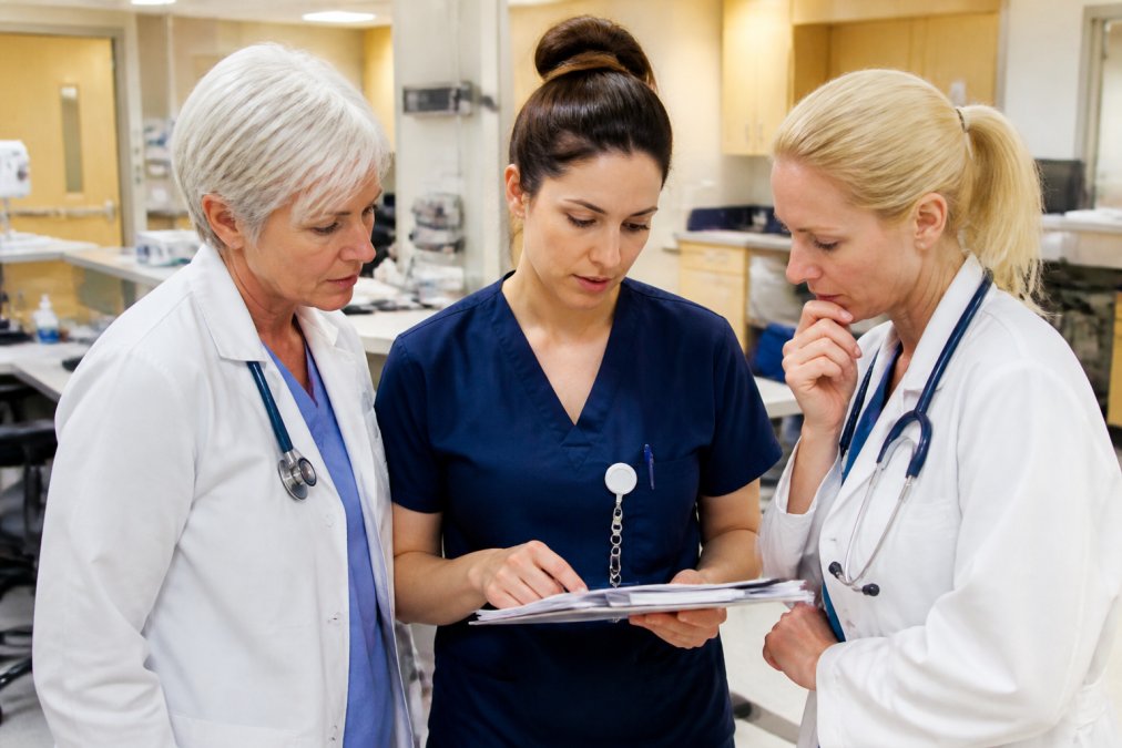 A nurse in navy scrubs consulting with two colleagues at a bright hospital workstation, reviewing a patient chart together with focused, thoughtful expressions