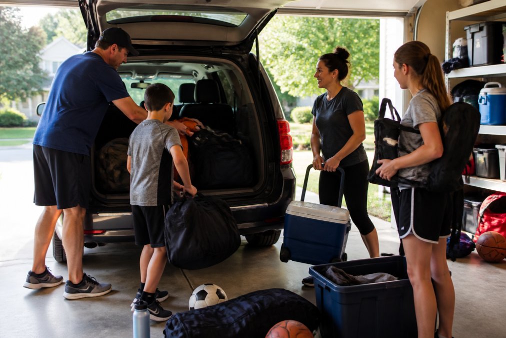 Family loading sports equipment and luggage into a minivan before heading to a tournament, with parents and children working together to pack efficiently, natural daylight streaming through the garage