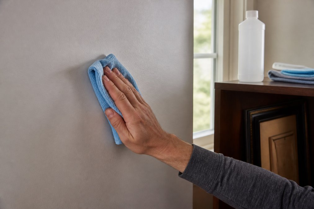 Close-up of hands wiping a clean painted interior wall with a microfiber cloth, a bottle of isopropyl alcohol on a nearby shelf, soft daylight from a window