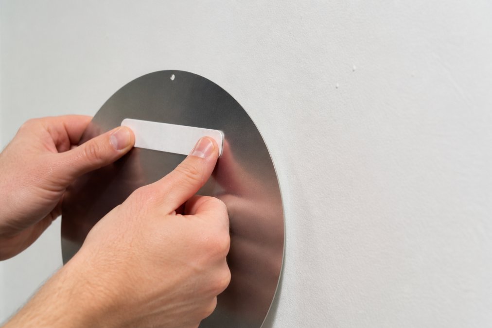 Close-up of a person's hands pressing a heavy-duty adhesive strip firmly onto the back of a brushed aluminum sign, with a clean white drywall surface visible in the background, bright natural window light illuminating the workspace