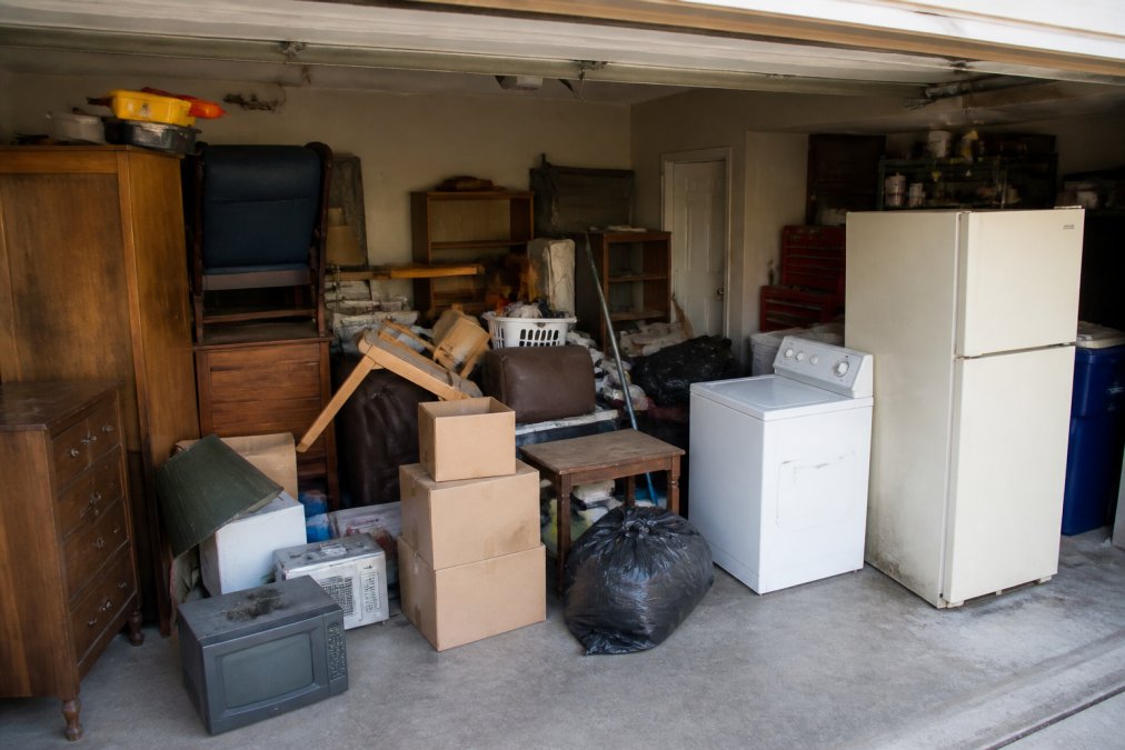 Cluttered garage with stacked furniture, old appliances, boxes, and miscellaneous items awaiting removal, showing typical before state of a residential garage cleanout project