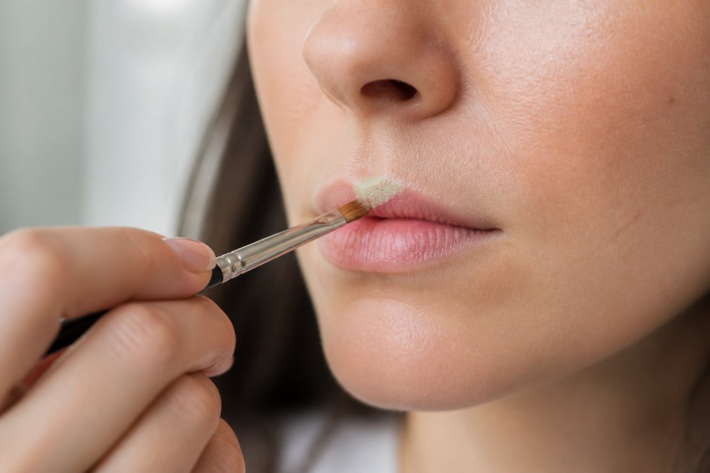 Close-up of hands applying green color-correcting concealer to a lip area with a fine makeup brush, showing precise blending technique with natural daylight