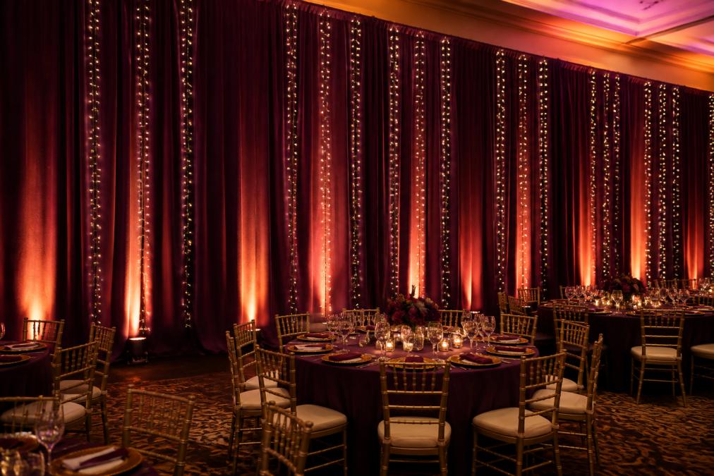 Professional event venue with warm uplighting illuminating draped walls in rich jewel tones, curtain lights strung across panels creating a glowing backdrop, showing how colored lighting transforms the aesthetic and creates depth in the space