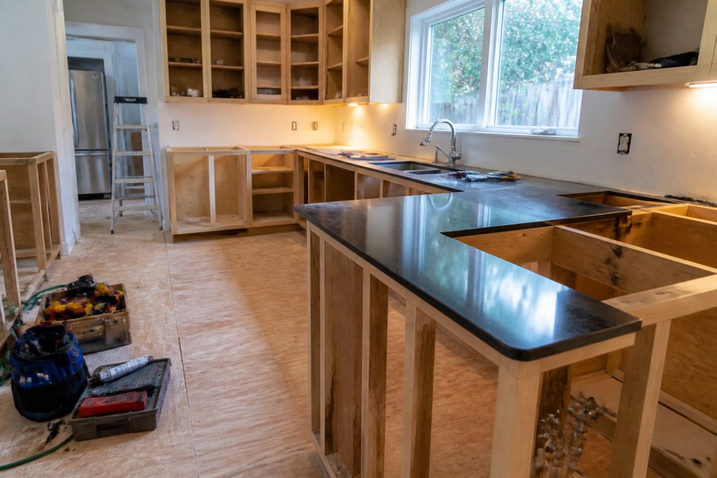 A kitchen mid-renovation showing exposed cabinetry framing and new countertop installation in progress, with tools organized neatly on a clean plywood-covered floor under bright work lighting