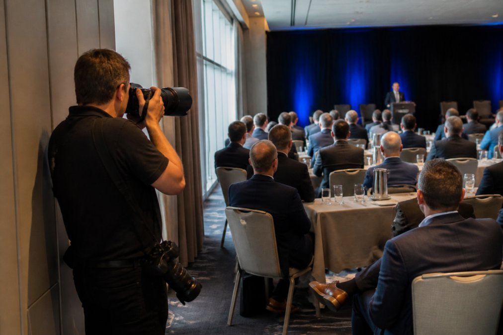 Event photographer positioned strategically on the sidelines of a conference session, capturing candid moments of speakers and attendees without being intrusive, with soft natural light from windows