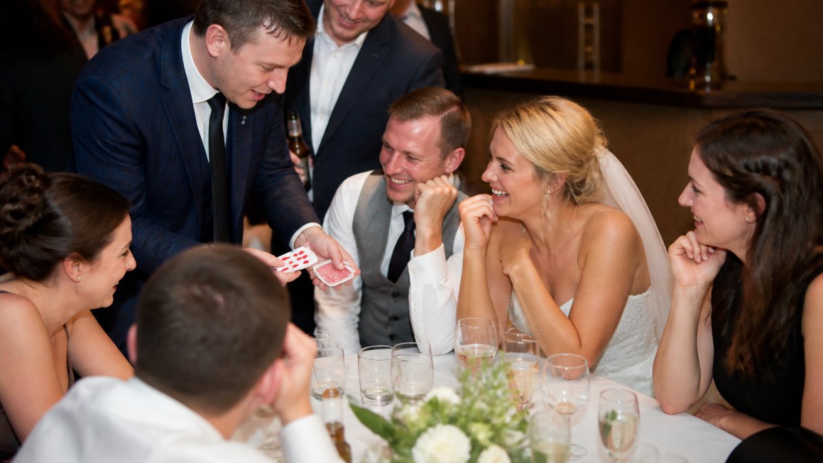 Professional magician performing close-up card magic at a wedding reception table, with guests gathered around smiling and engaged, warm ambient lighting on their faces
