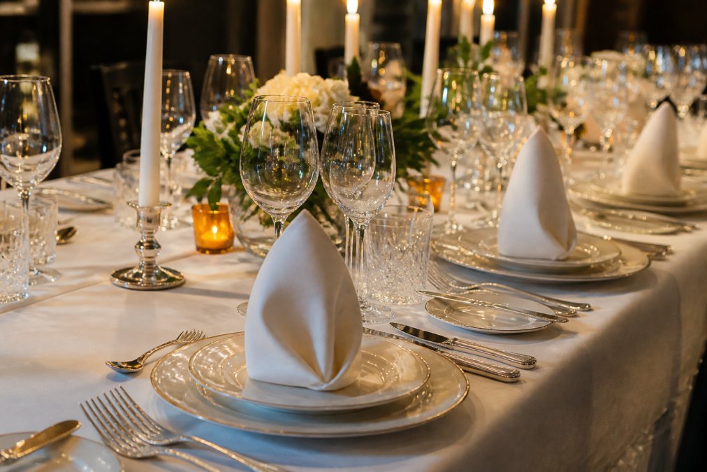 Elegantly set formal dining table with fine bone china plates, crystal wine glasses, polished silver cutlery arranged precisely, white cloth napkins folded formally, place cards positioned above each setting, candlelight from tall tapers reflecting off glassware