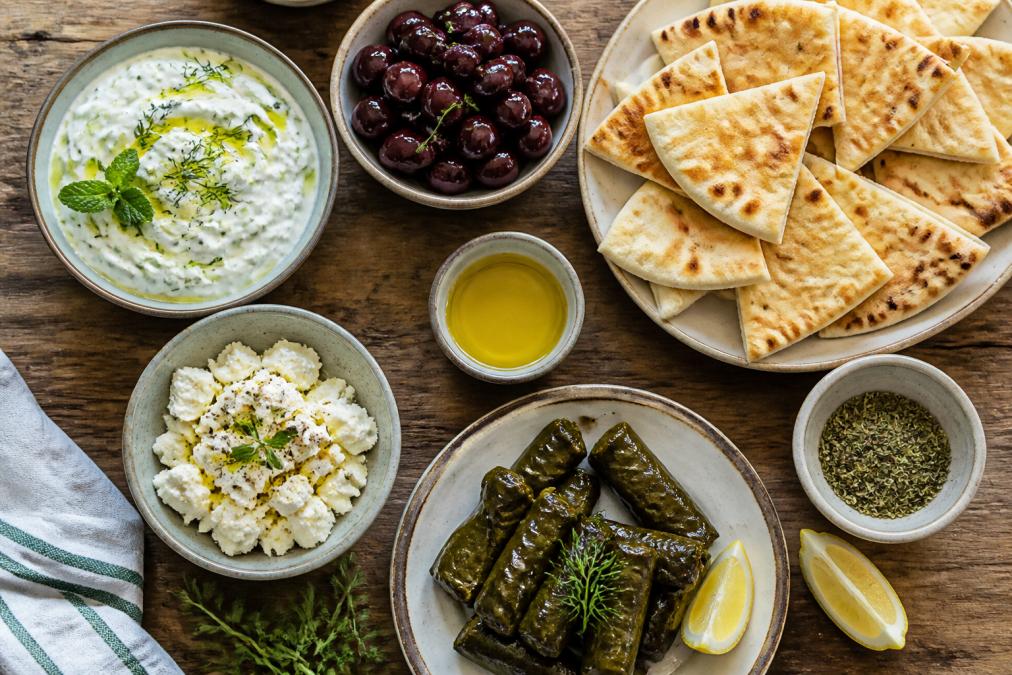 Overhead shot of a rustic wooden table filled with small sharing plates including tzatziki, feta, kalamata olives, dolmades, warm pita triangles, and a drizzle of extra virgin olive oil, fresh oregano sprigs and a lemon wedge, natural light, warm tones
