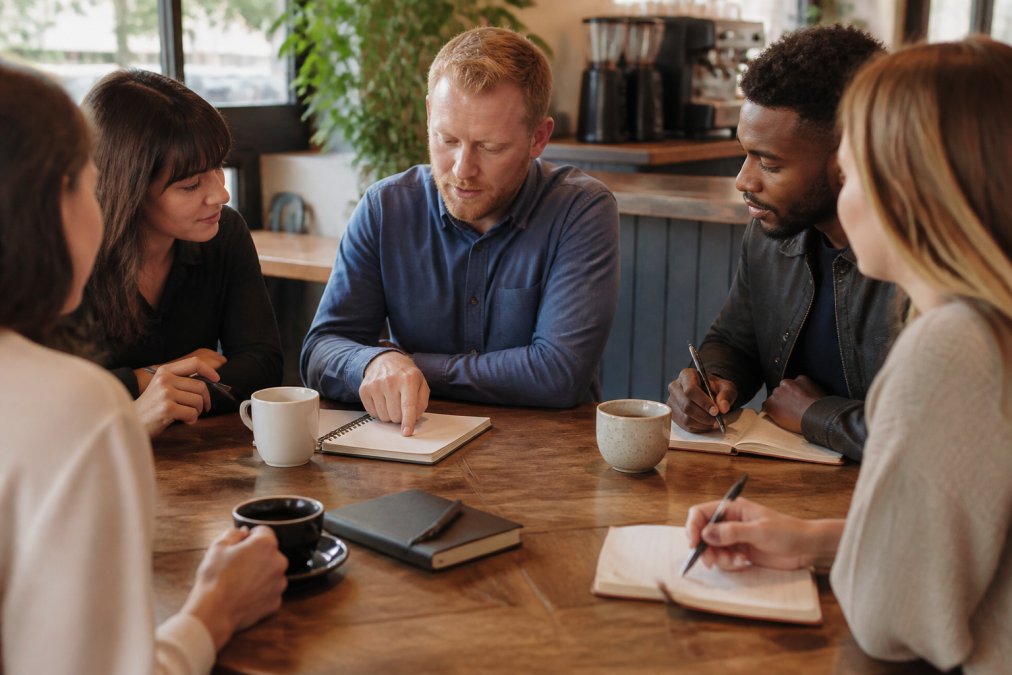 A small team gathered around a wooden table in a bright coffee shop meeting room, one person pointing at a notebook while others listen and take notes, coffee cups and laptops visible