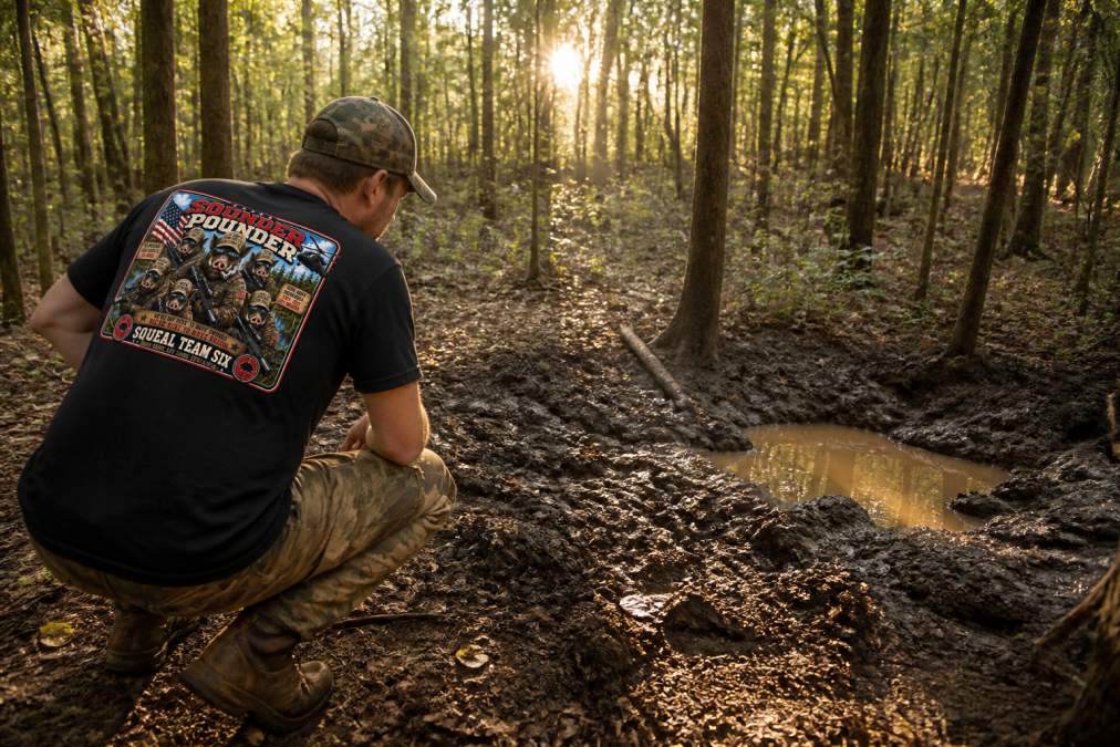 Hunter crouching to examine fresh rooting damage and muddy wallow sign on forest floor during daylight scouting, early morning light filtering through trees