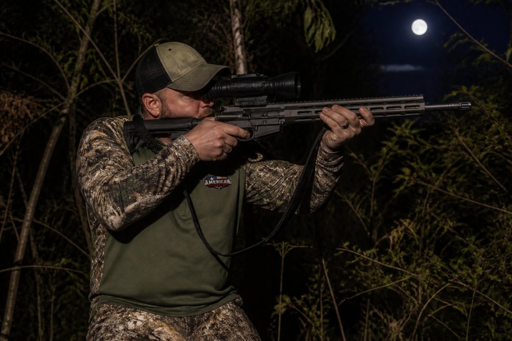 Hunter in camouflage using thermal optics equipment during nighttime hunt in wooded terrain with dense brush and moonlit sky