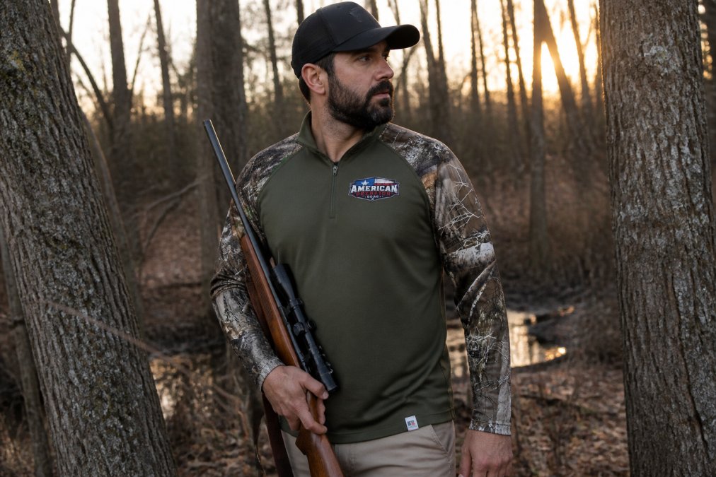 Outdoor hunter wearing moisture-wicking camouflage long sleeve shirt and holding rifle, positioned in natural wooded hunting environment at dusk