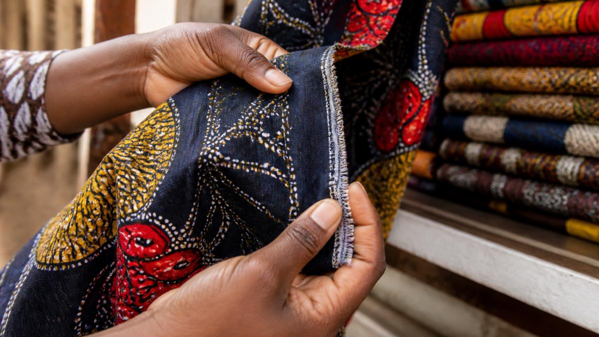 Close-up of hands examining authentic African print fabric, showing selvage edge with clear manufacturer markings, wax texture visible in the weave, and bold pattern details under natural daylight