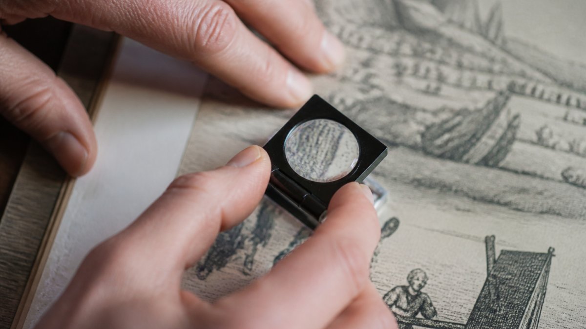 Close-up of hands holding a magnifying loupe over an antique print, examining fine dot patterns and ink lines of different printmaking techniques under magnification in natural light