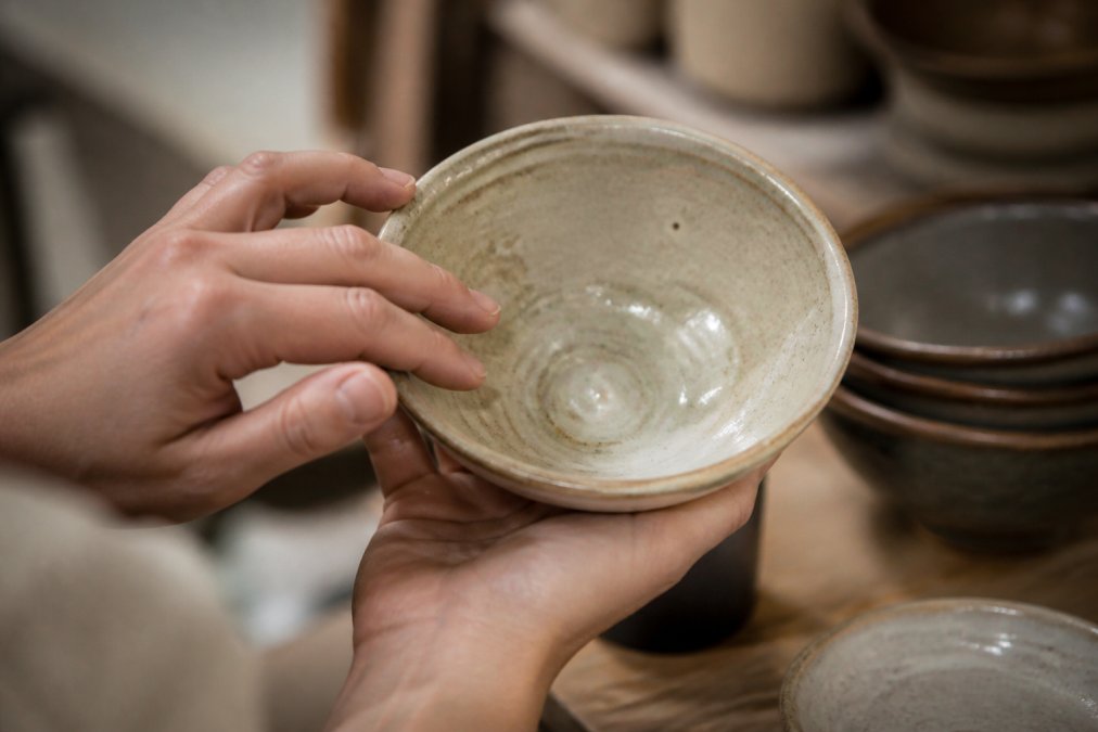 Close-up of hands examining the texture and finish of a hand-thrown ceramic bowl, showing subtle imperfections and glaze variations in warm natural light