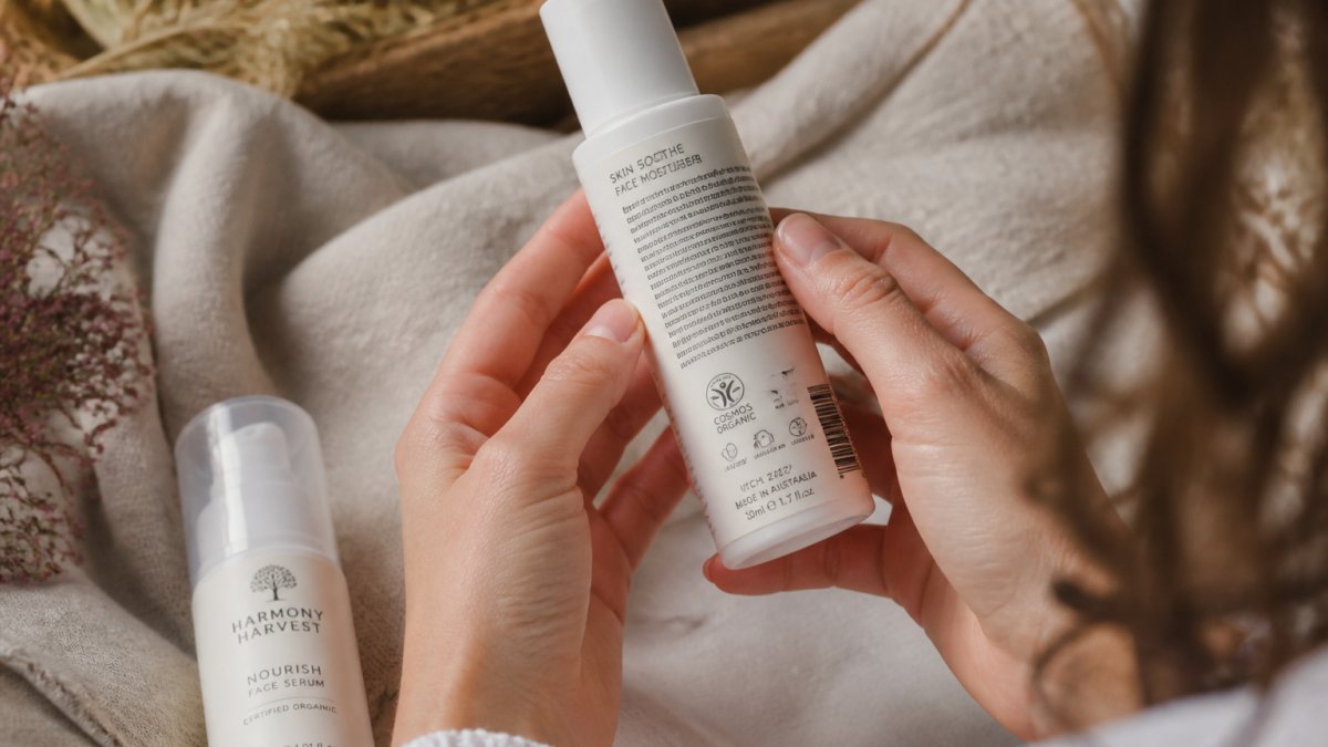 Close-up of a woman's hands turning a skincare bottle in soft natural light to read the certification logo, batch number and ingredient list on the label, with a second bottle resting on a linen surface nearby