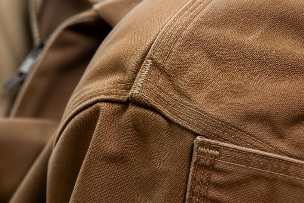 Close-up of expertly stitched seams on a heavy canvas work jacket, showing even, tight stitching with reinforced bar-tack patterns at stress points under natural daylight