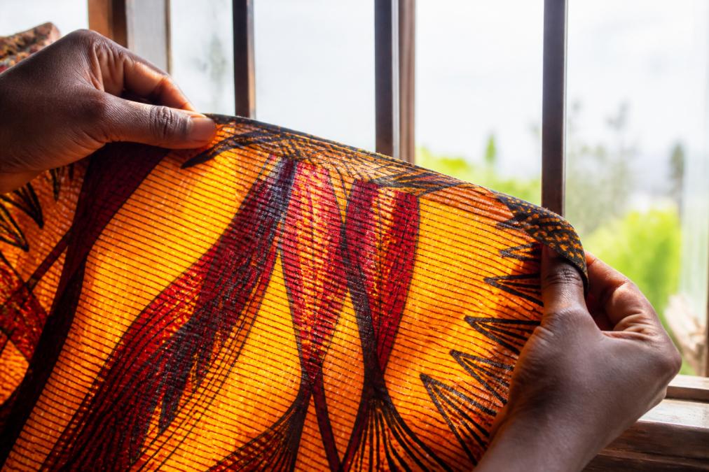 Close-up of hands holding a brightly patterned East African fabric up to a sunlit window, showing the vivid pattern visible through the cloth and the texture of the weave