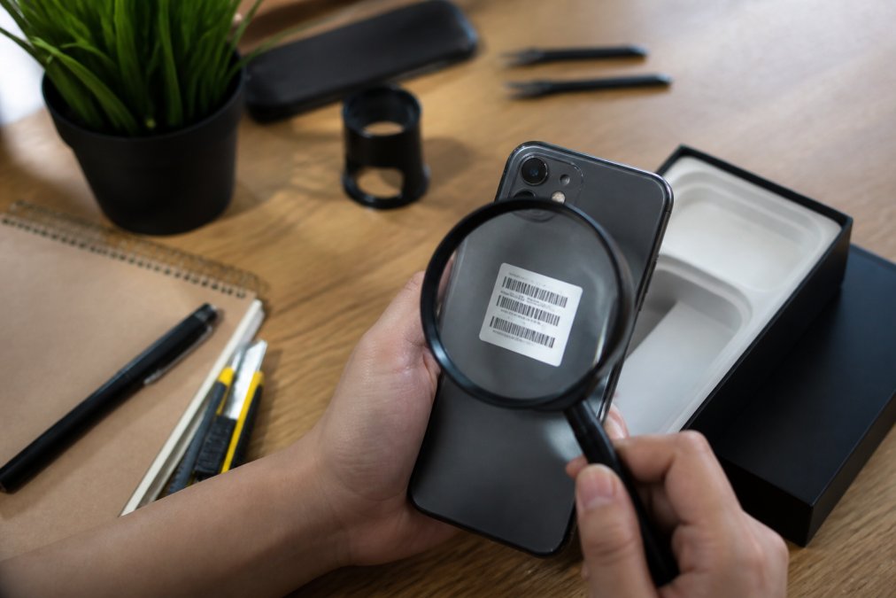 Hands holding a smartphone and original packaging while inspecting a serial number with a magnifying glass