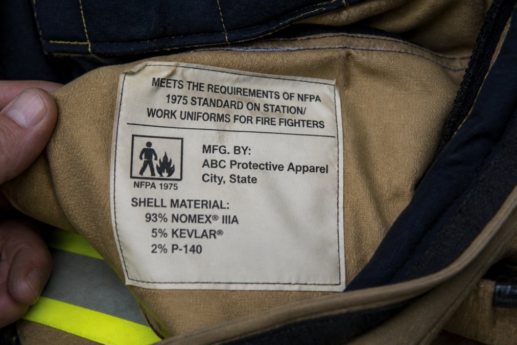 Close-up of a firefighter uniform label showing NFPA 1975 certification marking, manufacturer name, and fiber content clearly visible under natural lighting