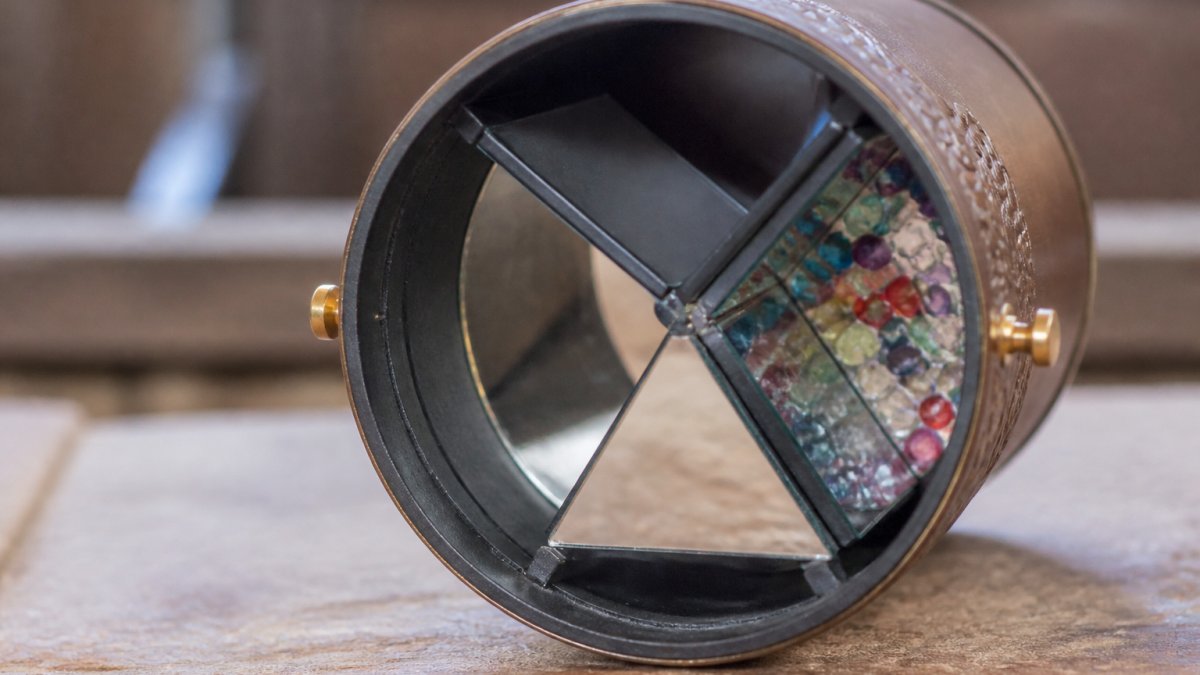Close-up of an open handcrafted kaleidoscope showing the interior three-mirror optical system with light reflecting off polished mirrors and the object chamber visible