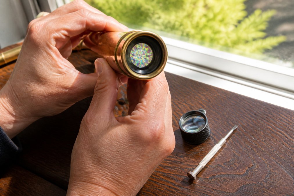 Close-up of hands holding a brass handmade kaleidoscope up to a window, light refracting through the eyepiece, with a loupe and small screwdriver on a wooden table nearby