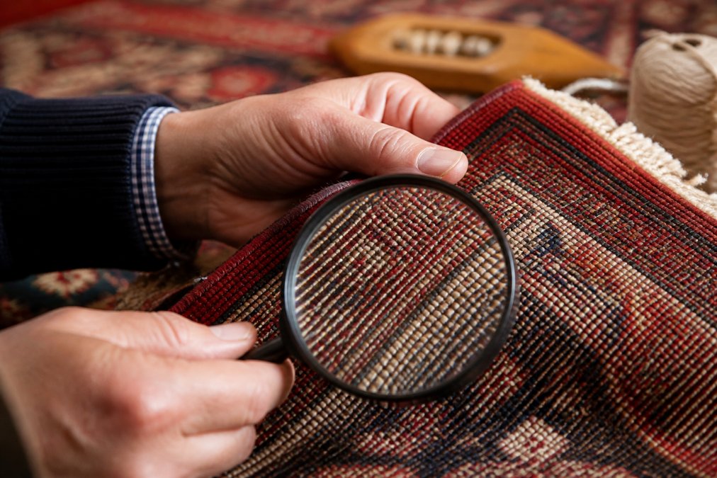 Close-up of hands holding a magnifying glass over the back of a Persian rug, examining the intricate knot structure and weave pattern in detail under natural lighting