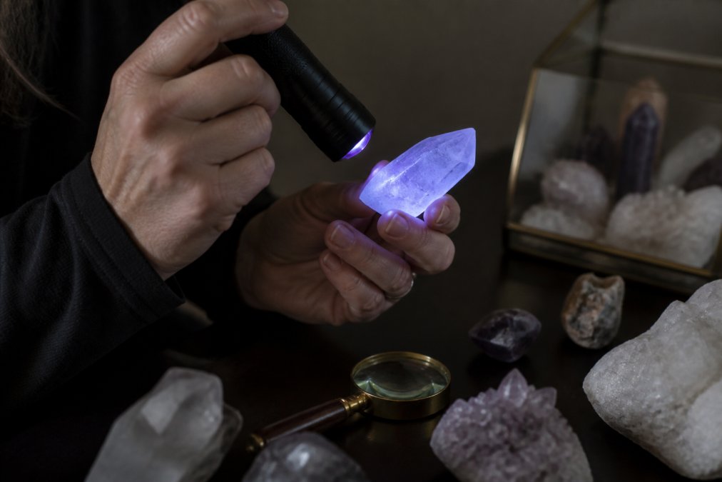 Hands holding a clear quartz crystal specimen up to a UV flashlight in a dimly lit room, showing a soft glow, with other crystals and a magnifying glass on the table nearby