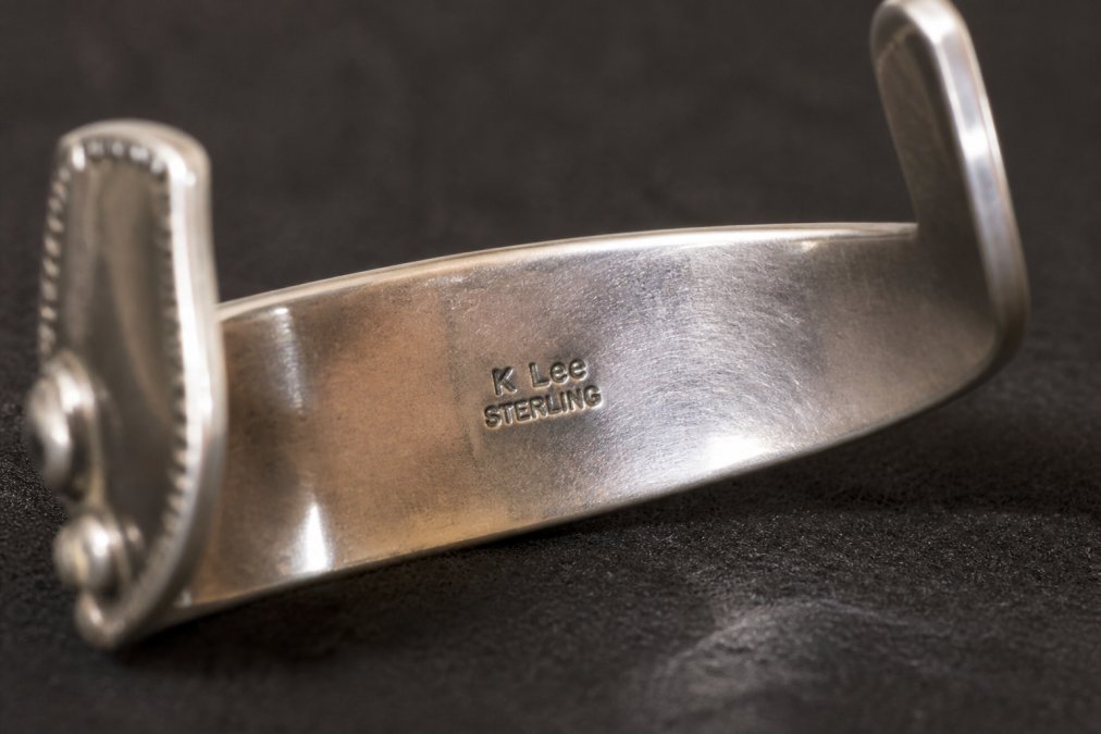 Extreme close-up of a silversmith's hallmark hand-stamped into the back of a sterling silver Navajo cuff bracelet, showing crisp maker's mark lettering and a sterling stamp against oxidized silver, photographed under bright directional lighting on a dark felt surface