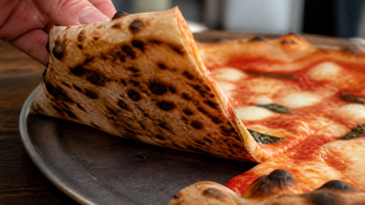 Close-up of a hand lifting a slice of pizza, showing a puffed, charred crust and a slightly crisp bottom with visible leopard spotting