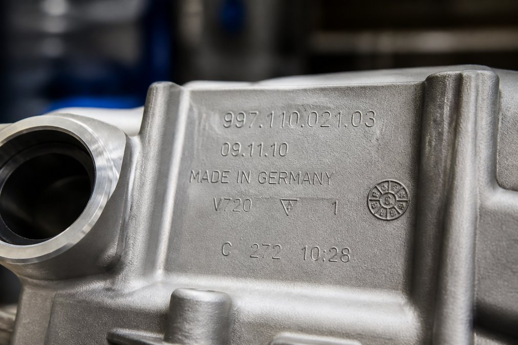 Close-up of a Porsche OEM part with clearly stamped part number, manufacturing date code, and quality control markings visible on brushed metal surface under bright halogen lighting