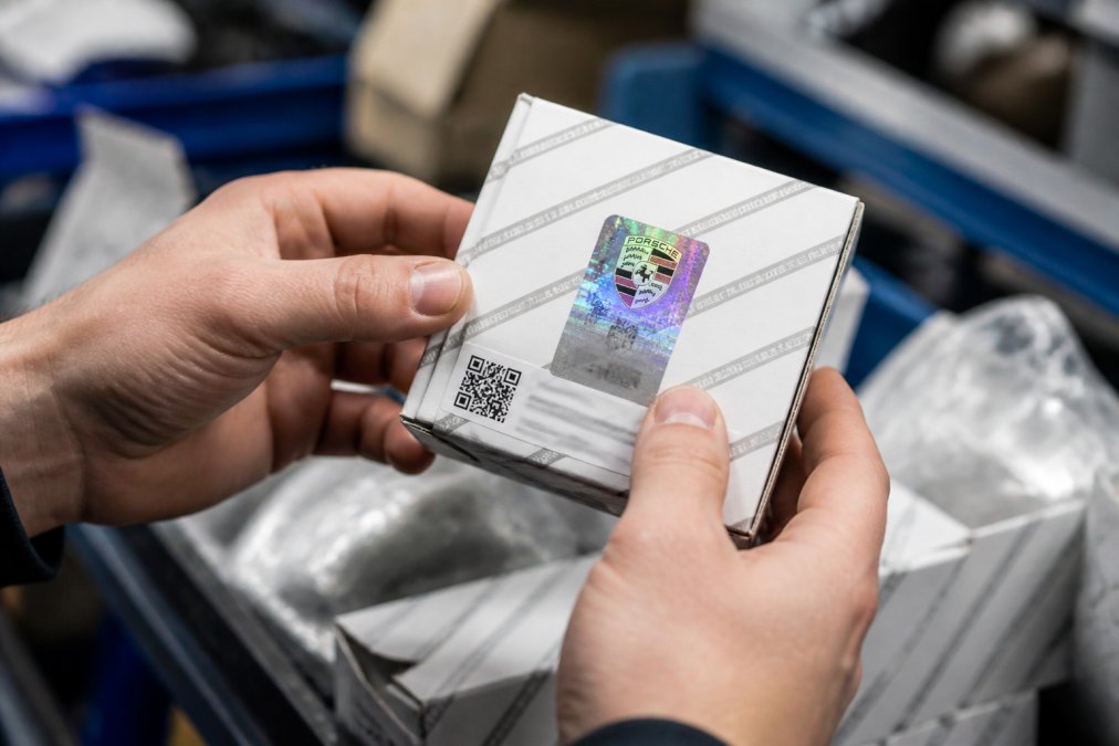 Hands holding a genuine Porsche OEM part box displaying multi-layered holographic security labels with color-shifting effects, official Porsche branding in crisp printing, batch tracking codes, and sealed protective packaging under studio lighting