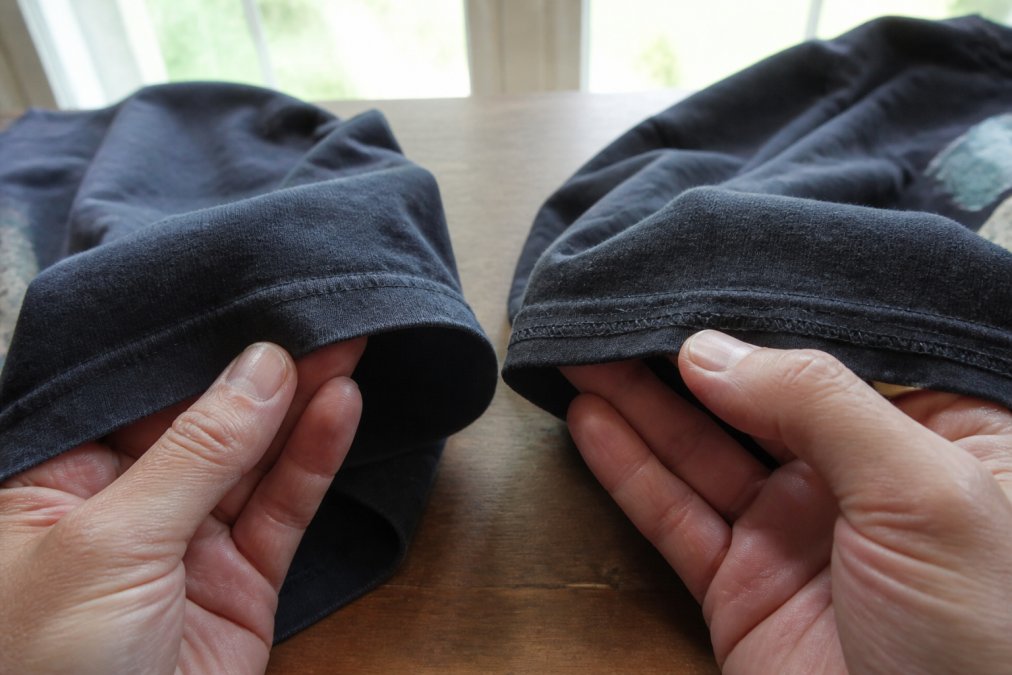 A collector's hands holding the hem of a faded vintage t-shirt, clearly showing a single line of stitching along the bottom hem and sleeve opening, natural window light