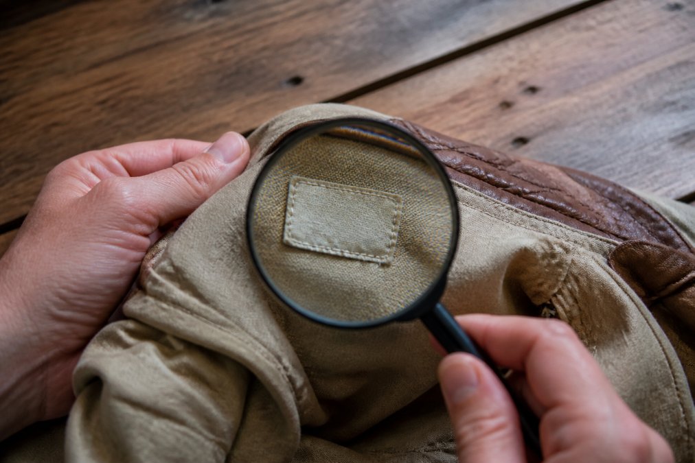 Close-up of hands holding a magnifying glass over an aged clothing tag inside a vintage jacket, with a wooden table and soft natural light in the background